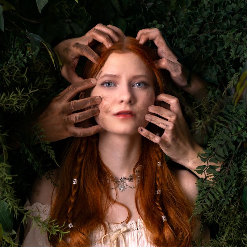A woman's face is surrounded by leaves and ivy with four male hands forcefully grabbing her face and hair.