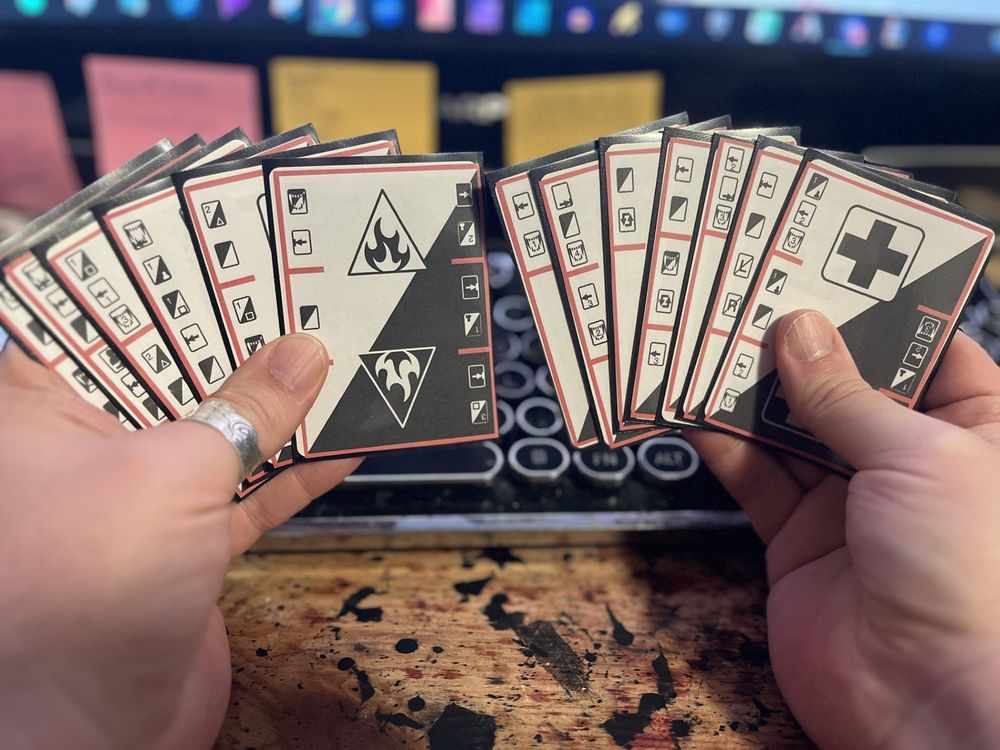 Photo of my hands holding 13 cards, 6 in one hand, 7 in the other. Each card has a light and dark side, with symbols denoting the ship room and instructions for how to play. 