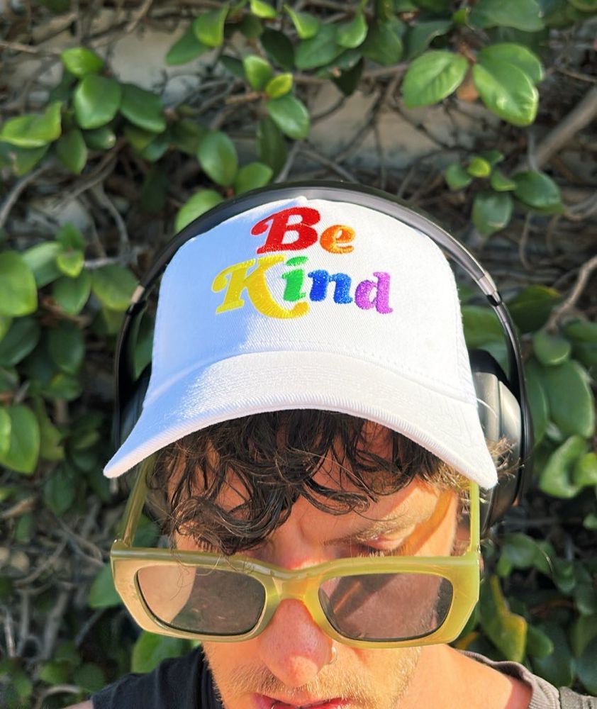 Selfie of Kasey against a greenery-covered wall, wearing a white baseball cap that says “Be Kind” in rainbow lettering 🖤 SH!NER are a queer alternative rock band in LA