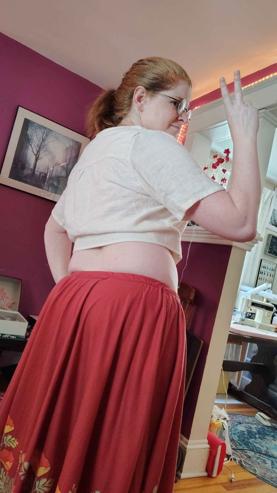 a shot of me from behind, standing and throwing up a peace sign. you can see the yoke of the shirt and the substantial crop showing my midsection. i'm also wearing a red skirt with flowers.