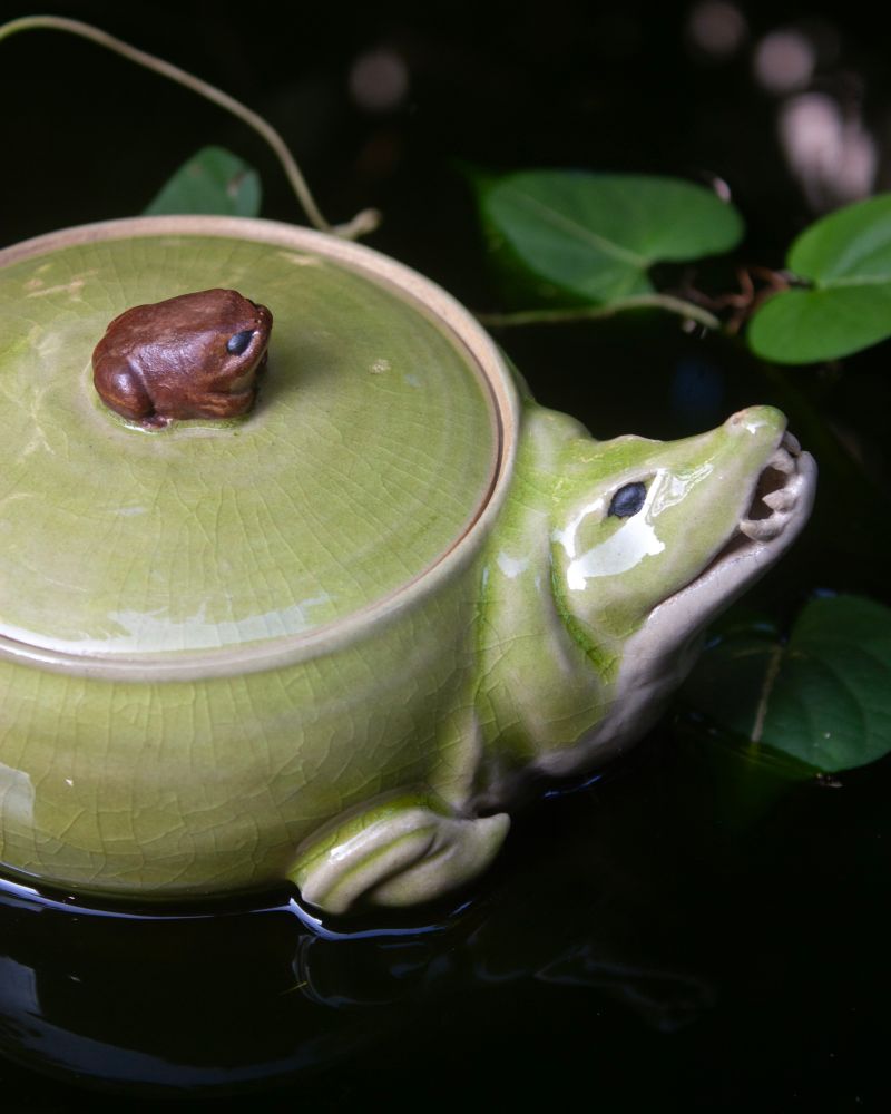 Close up of chubby ceramic crocodile jar face and lid showcasing detail on green crackle glaze and brown texture of small toad on top.
