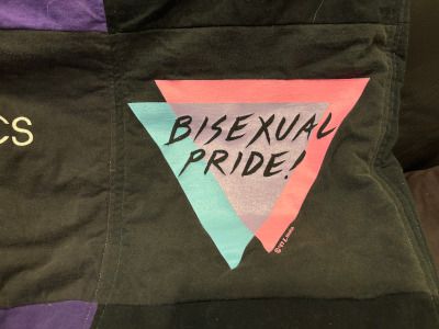 A section of a quilt made from T-shirts. This square says Bisexual Pride superimposed on two triangles, one pink and one blue, that overlap to make a purple triangle, which was a popular bisexuality symbol in the 1990s. (It seems to be less popular now, though other variations on the pink-purple-blue are around.) 
