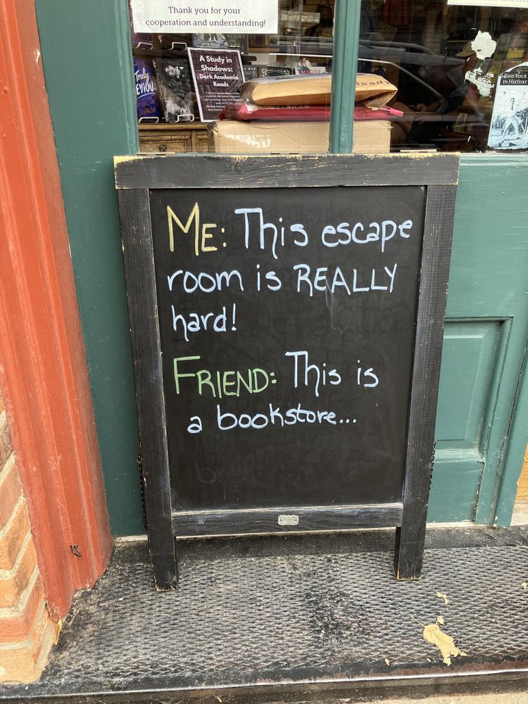Chalkboard sign outside a retail establishment. Dialogue: Me: This escape room is really hard! Friend: this is a bookstore…
