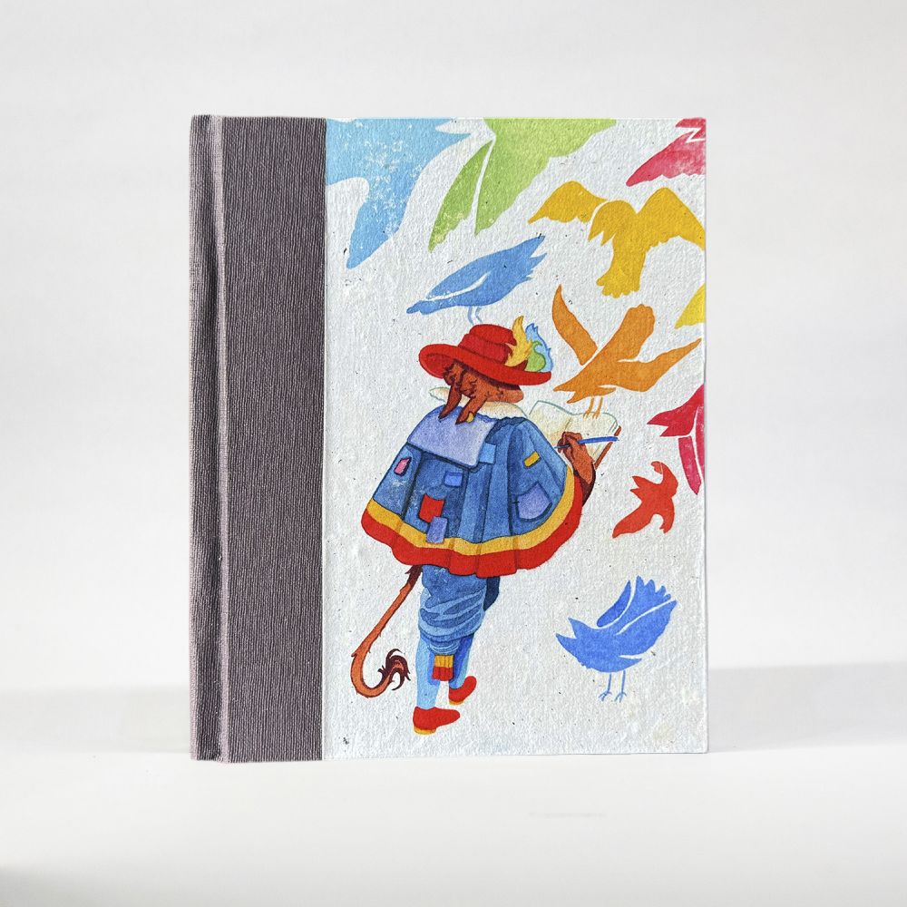 A notebook with the cover visible stands on a white background. The spine is a muted purple paper, with a natural white, textured, handmade paper cover featuring an image of a brown, fuzzy creature in a patchwork cloak surrounded by colorful birds. The creature writes in a journal as they walk.