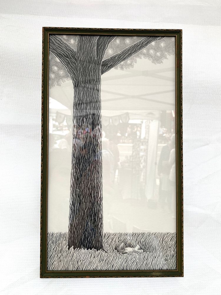 A photograph of a framed black and white drawing of an acorn kid lying in the grass beneath a tall oak tree. In the glass, reflections of fair patrons can be seen.