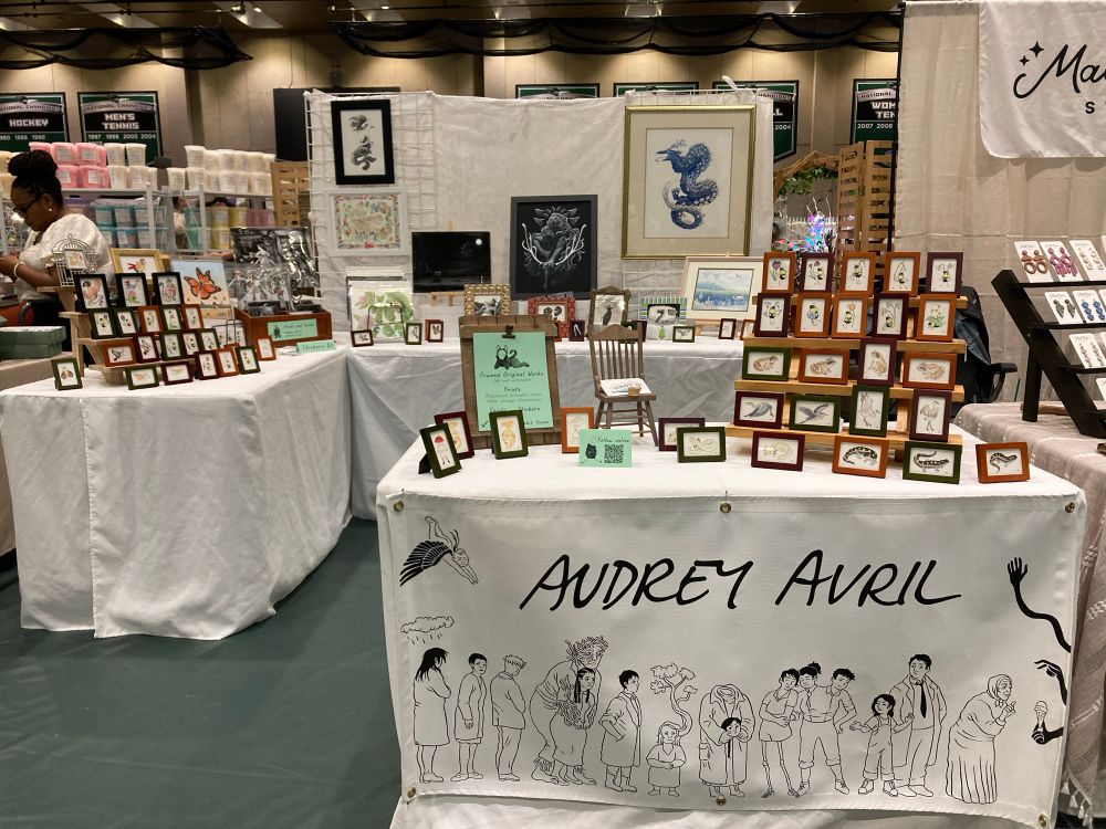 A photograph of an artist’s booth, featuring small frames on a table with banner in the foreground, and an assortment of small and large frames on the tables and wall in the background.