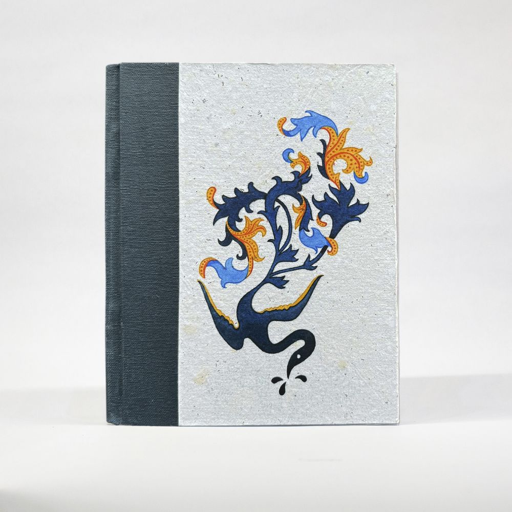 A notebook with the cover visible stands on a white background. The spine is a muted blue paper, with a natural white, textured, handmade paper cover featuring an image of a abstracted bird creature, its tail branching off into interwoven leaves of navy, light blue, and yellow.