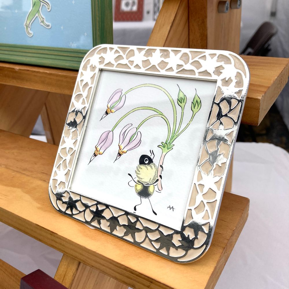 An ink and watercolor illustration in a patterned metal frame displaying a bee and a stalk of a shooting star plant.