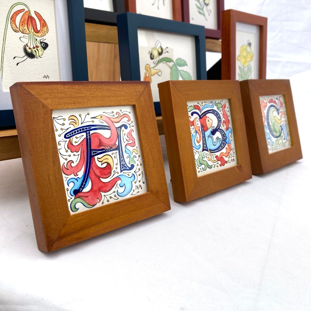 A photograph of small framed ink and watercolor art. In the foreground in three brown wood frames are three historiated capital letters done in the style of medieval manuscript illustration.