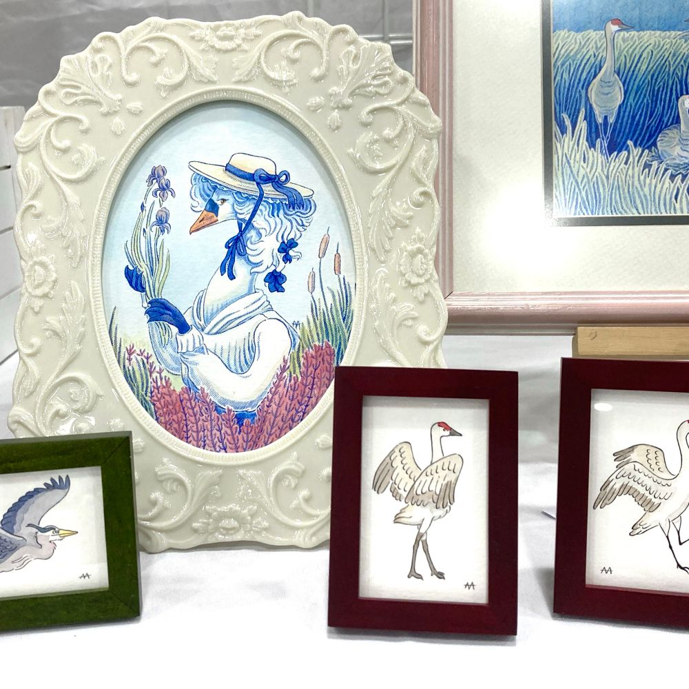 A collection of framed ink and watercolor artworks on a bright white tablecloth with a white backdrop. Three small frames feature two sandhill cranes and a great gray heron, strutting, flying, and stretching their wings. A larger, white scroll-work frame with a circular opening features a graceful swan in a white dress, rococo wig and hat in a colorful floral backdrop. The corner of a pink-framed piece shows a part of a larger grassland composition with a sandhill crane among the waving field. 