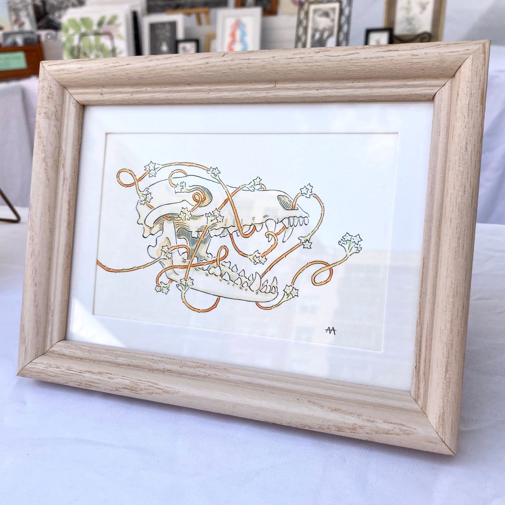 A photograph of a framed ink and watercolor illustration. In the artwork, a flowering orange dodder plant wraps around a coyote skull.