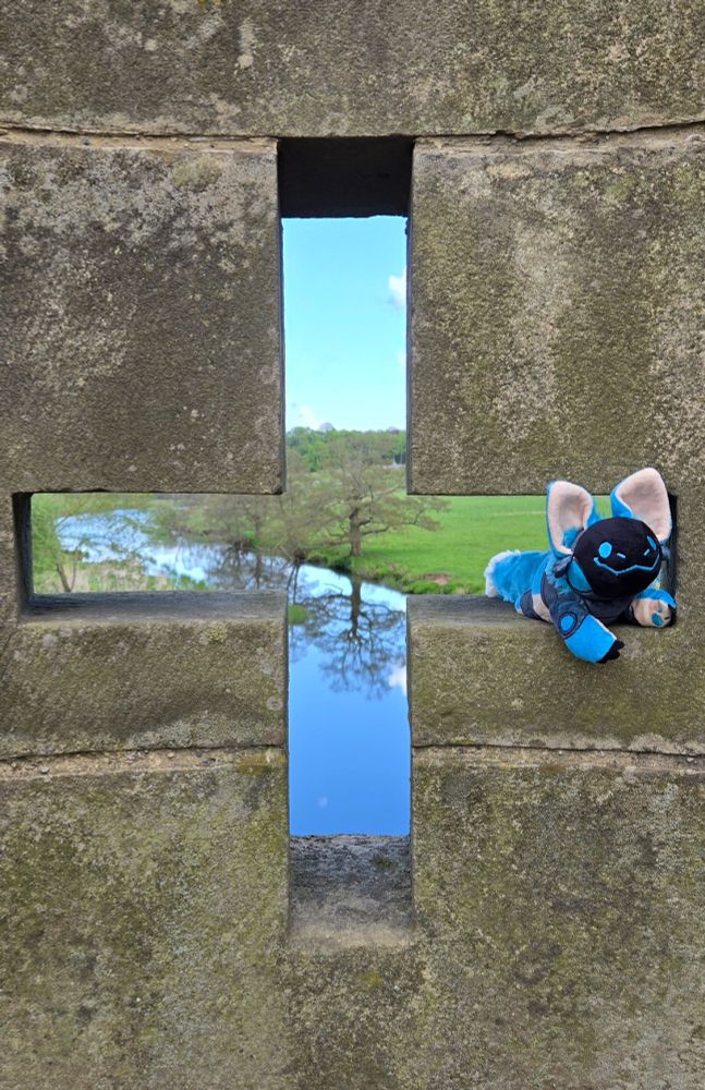 A smiling blue protobean is perched in the right arm of a cross-shaped cutout in an stone wall. A blue sky, a green grass field, a blue stream, and a few trees and their reflections in the water are visible through the cutout. 