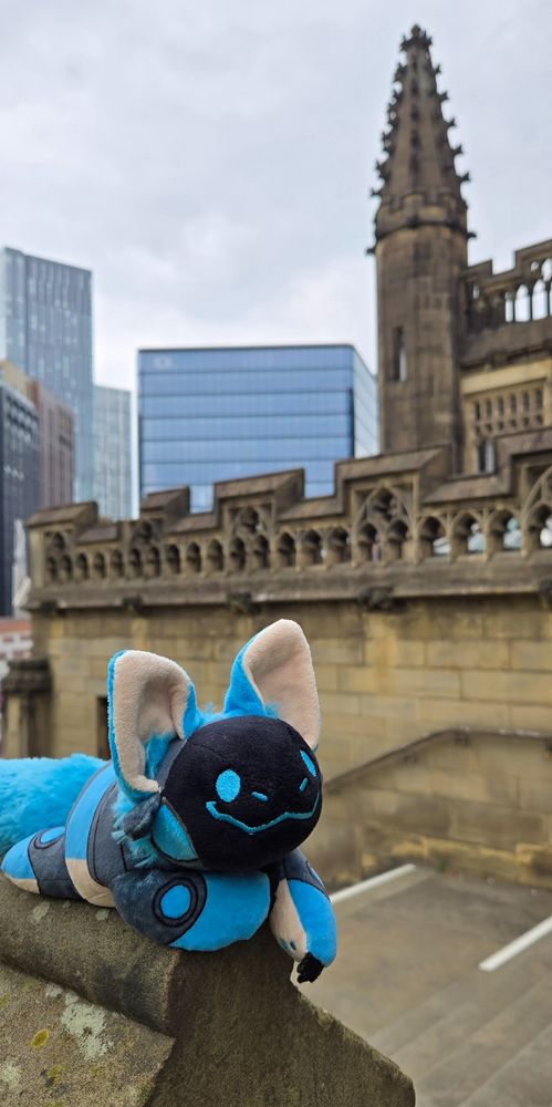 A smiling blue protobean is perched on a stone railing of a stone staircase. Behind him is an ornate stone cathedral wall and spire, and behind that are glass skyscrapers and a grey sky.