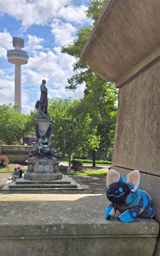 A smiling blue protobean is sitting on a stone wall overlooking the William Gladstone statue in St. John's garden. In the distant background, the Radio City tower rises out of a line of green-leaved trees against a blue sky full of  puffy white clouds.