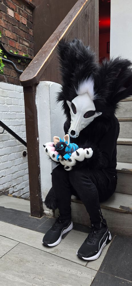 A skulldog fursuiter with black and white fur is sitting on some stairs looking down at a smiling blue protobean held in his paws. He's wearing all black clothes with black and white shoes.