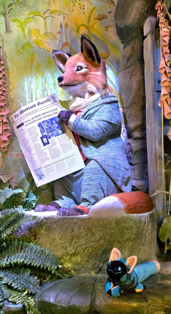 A smiling blue protobean is sitting on a stone wall beneath a rock. On top of the rock, a realistic fox puppet is sitting and reading a newspaper. The fox is wearing a gray suit and white scarf. The newspaper headline reads "LOCAL AUTHOR BRINGS FAME TO WOODLAND CHARACTERS"