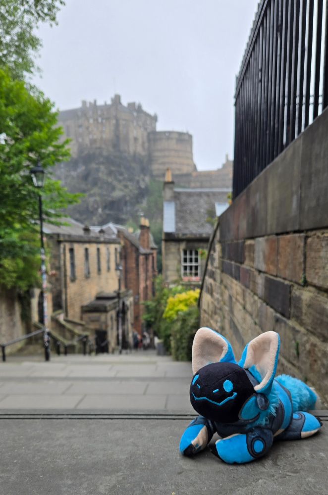 A smiling blue protobean is perched at the top of an outdoor stone staircase leading down into the background. Stone buildings, trees and shrubs line the way down. In the distant background, Edinburgh Castle is visible on top of its hill against a pale grey sky.
