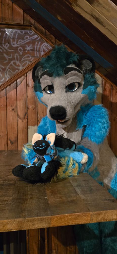 A smiling blue protobean being held in a blue and gray wolf-otter's paws. The wotter is sitting at a wooden table against a wood-panel wall.