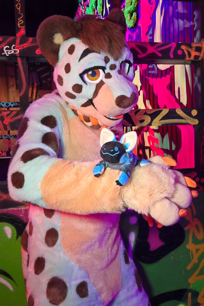A cheetah fursuiter is standing against a vibrantly colored graffiti background. His fur is mint green and cream with brown spots. His forearm is held across his body, and a smiling blue protobean is perched on it.