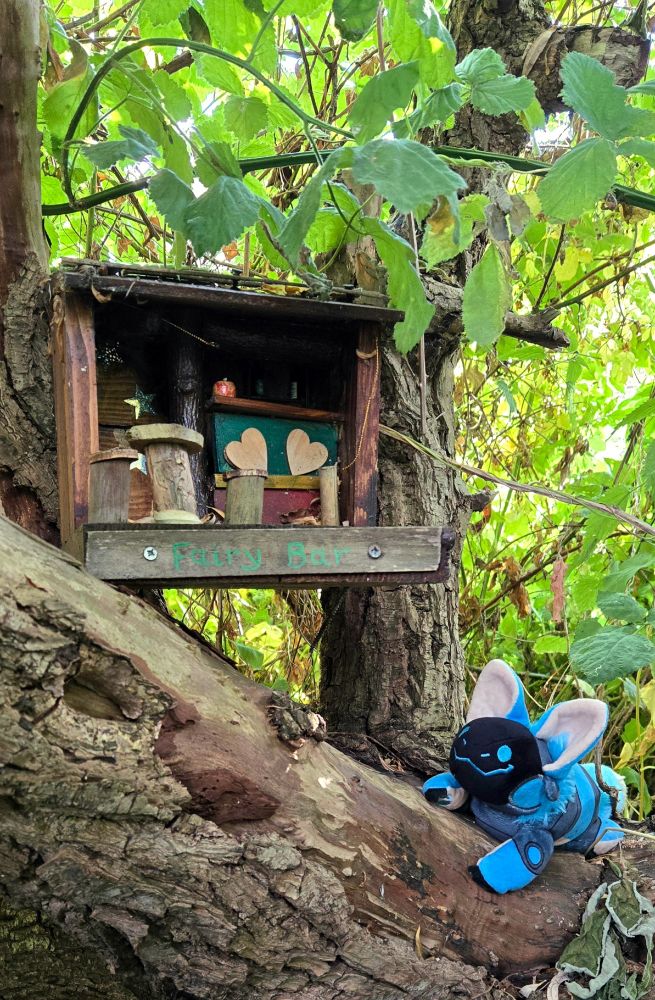 A small wooden tree house is attached between two tree trunks with a larger trunk sloping through the foreground. There are green leaves and bits of sunlight shining behind it. The tree house is square and has a small countertop and four mismatched wooden bar stools. On the front panel, "Fairy Bar" is written in green letters. A smiling blue protobean is perched on the trunk beneath it.