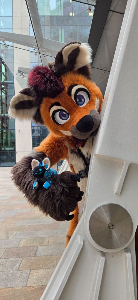 An orange maned wolf fursuiter with a dark red mohawk is leaning out from behind an angled white metal support beam. A smiling blue protobean is perched on his arm. Behind him, a stone-paved plaza leads to tall buildings with glass windows. 