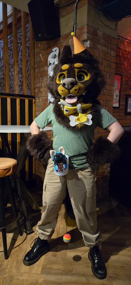 A smiling blue protobean hanging out of a cup tied around a partial fursuiter's waist. The suiter is wearing a brown and tan dog head with an orange party hat on one ear, brown furry paws, a big plush yellow collar with a bone-shaped name tag, a green shirt, and khaki pants.