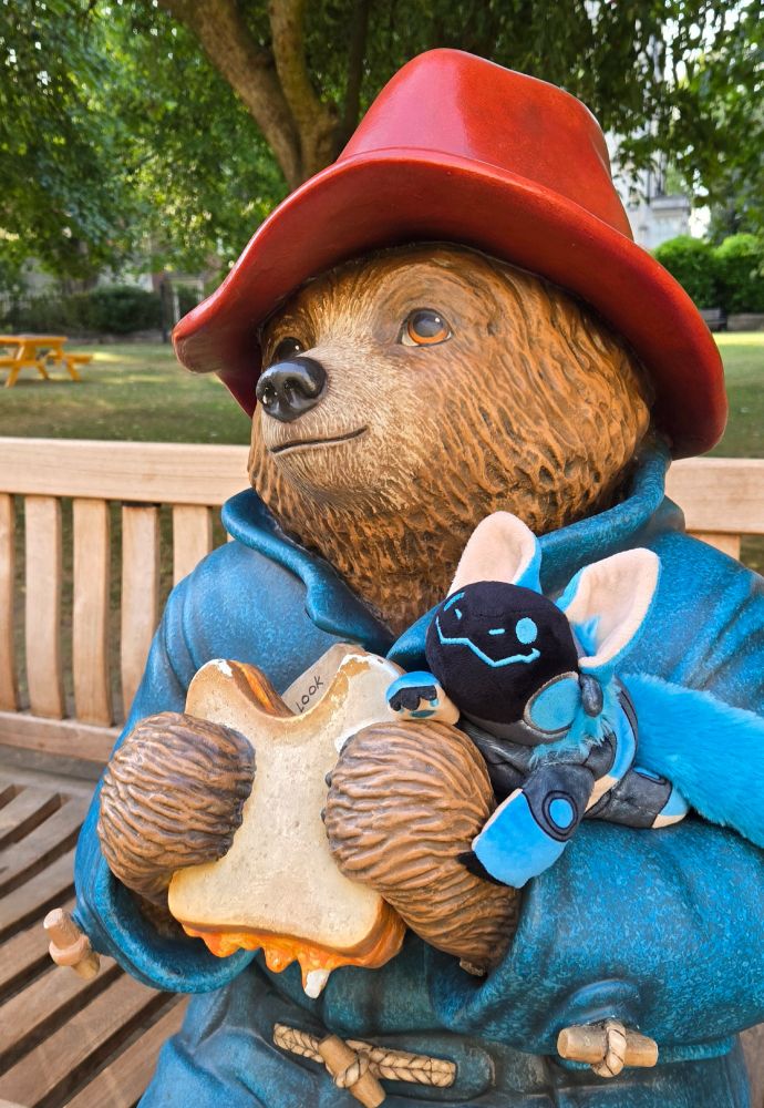 A smiling blue protobean is perched on the arm of a Paddington Bear statue seated on a wooden bench in a park. Paddington Bear is wearing a red hat and a blue anorak coat, and is holding a marmalade sandwich with one bite taken out of it. He is looking up and smiling, gazing wistfully into the distance. Behind them, there is a lawn of green grass, several leafy trees, and a wooden picnic table.