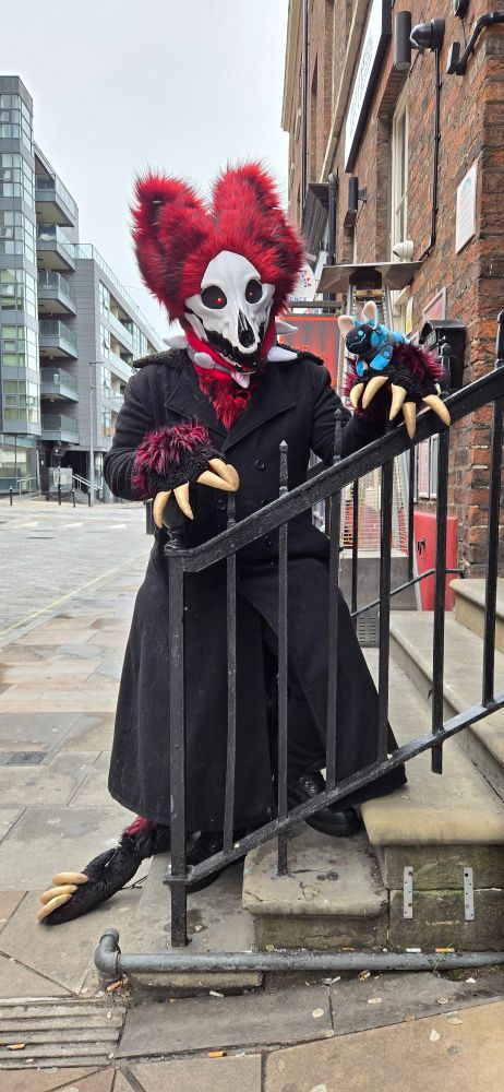A skull dog fursuiter with dark red fur and a white canine skull face is standing behind a black metal railing on a concrete stoop. His large-clawed paws are resting on top of the railing, and on one paw a smiling blue protobean is perched. He is wearing a long black coat. Behind him, a city street stretches to a pale grey sky with modern glass buildings on one side and older brick buildings on the other. 