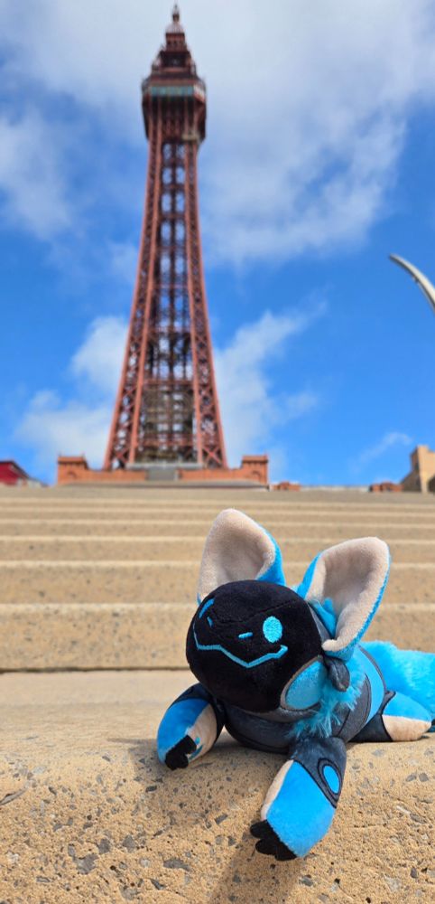 A smiling blue protobean is sitting on tan cement steps in the sunshine. At the top of the steps, the red metal Blackpool Tower is visible against a blue sky with puffy white clouds.