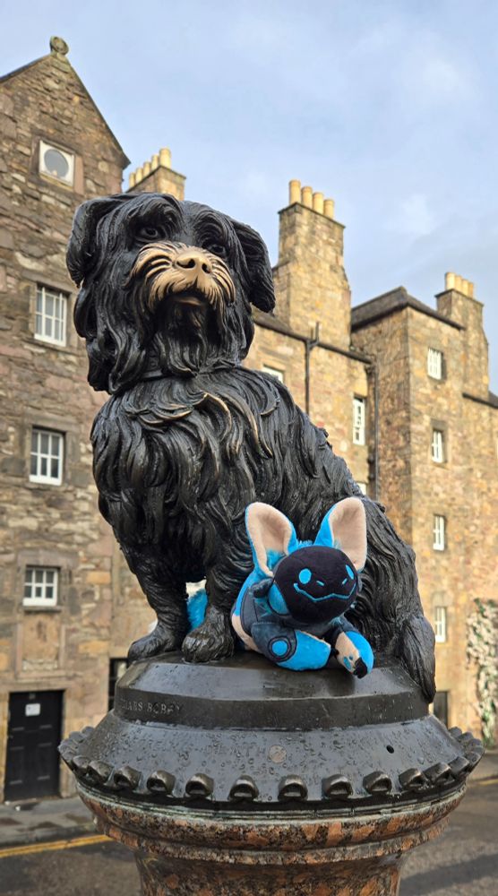 A smiling blue protobean is wedged between the legs of the statue of Greyfriars Bobby, a dark terrier statue with a gold nose where tourists have rubbed it. Behind them, stone buildings and chimneys rise against a cloudy yet sunny early morning sky.