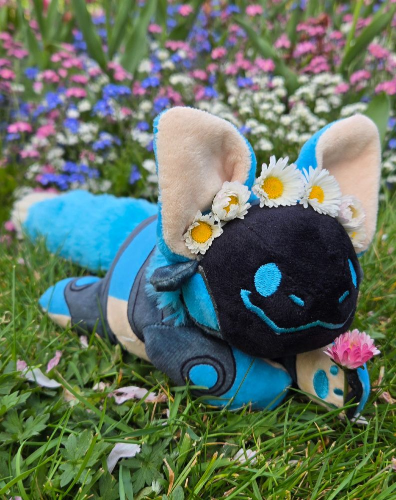 A smiling blue protobean laying on the grass against a backdrop of pink, blue, and white flowers. He is wearing a crown of white daisies and holding a pink clover blossom in his paw.