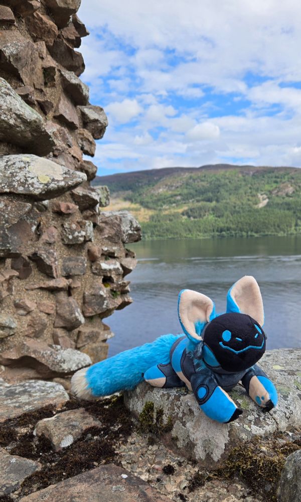 A smiling blue protobean is perched on a stone castle wall, which rises up on the left side of the photo. In the middle and right side of the photo, the blue-green water of Loch Ness and the hills across the shore are in the background. It is a sunny day, but the blue sky is full of white clouds.