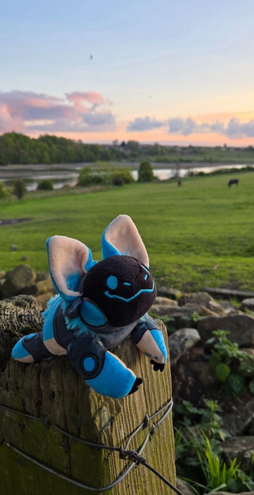 A smiling blue protobean sitting on a wooden fencepost, softly illuminated by the setting sun. Behind him is a grassy green hill with a couple of grazing horses, a shining river, and a pastel sunset in blue and orange, with a dusting of purple clouds.