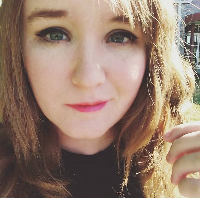 Elle, a feminine presenting nonbinary person with blue eyes and red hair, hair down, and smiling at the camera.
