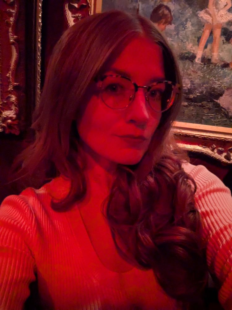 selfie of me under red light, my hair is curled, I am almost smiling, I am wearing glasses.