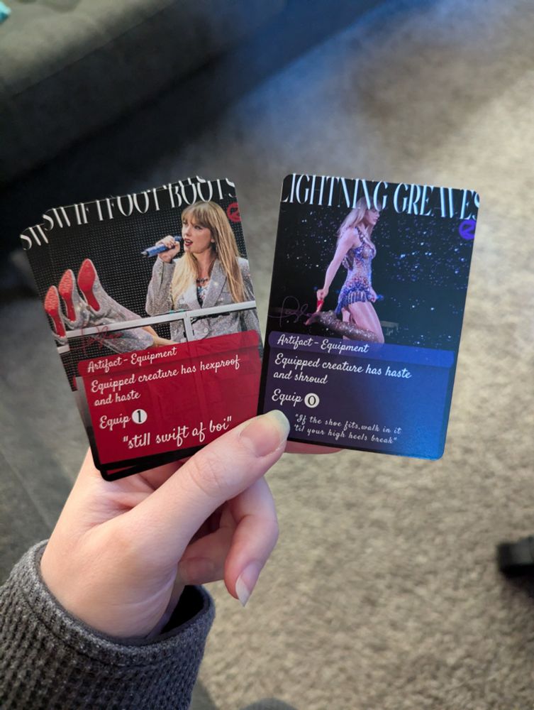 my hand holding alters of magic the gathering cards
swiftfoot boots shows Taylor performing The Man at the Eras tour in KC (I think night 1), with her feet up on the desk. The flavor text is "still swift AF boi"

the other is lightning greaves and shows Taylor Swift after her heel broke while performing Cruel Summer during the Eras tour in Brazil (night 3). She's in the middle of removing the heel from the boot in the photo. The flavor text is "if the shoe fits walk in it till your high heels break."