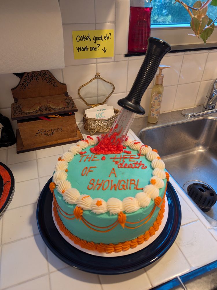 a gorgeous cake with mint/light blue frosting and orange accents
in a script matching the album font, it says "THE LIFE OF A SHOWGIRL" with "life" crossed out and "death" written in cursive below it.
there's a prop knife sticking out of the cake with red frosting blood splatter around it 