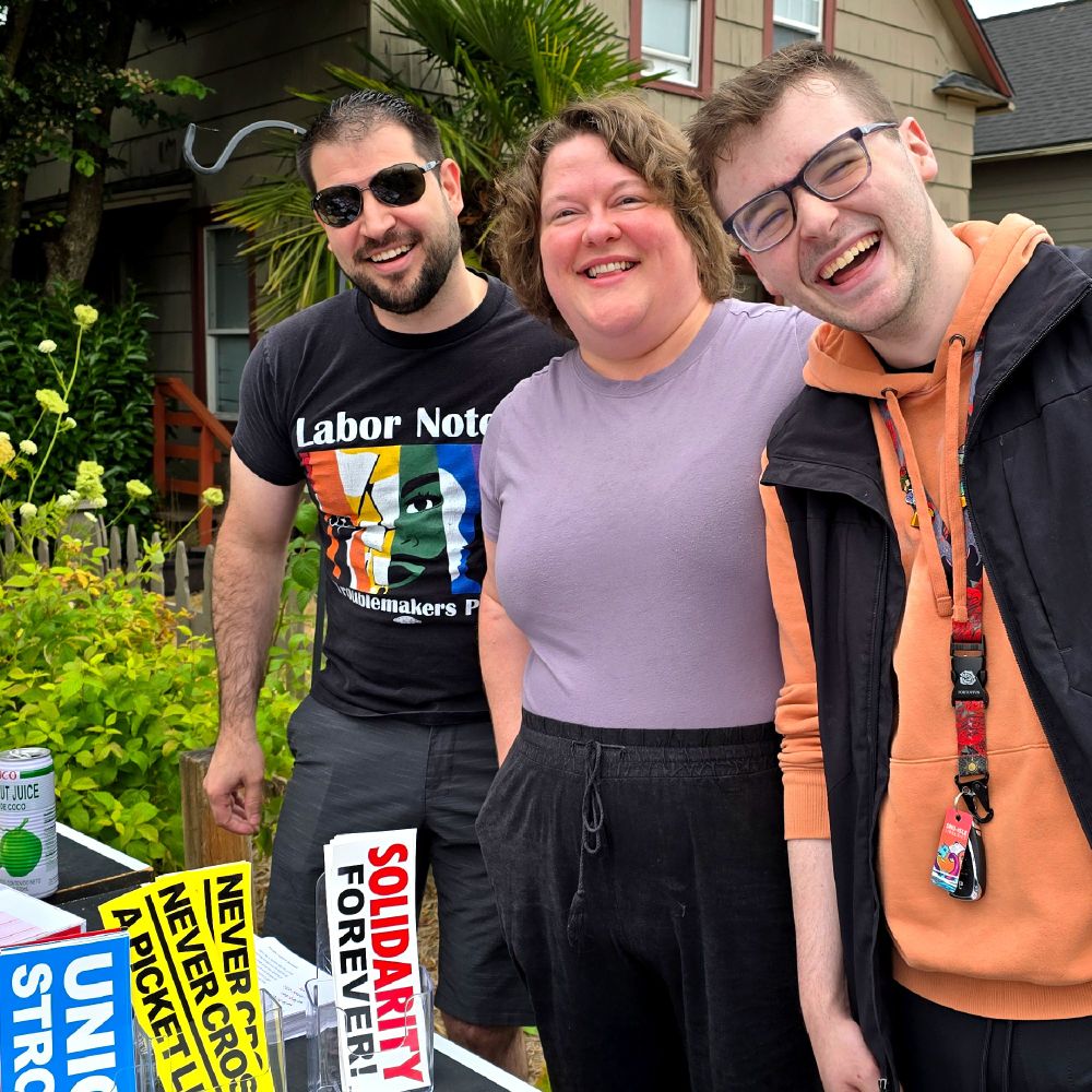 Three SnoCo DSA members, one of them an elected delegate going to Chicago, smile at the Hot Socialist Garage Sale. In front of them is a table with stickers that say “Solidarity Forever, Union Strong and Never Cross a Picket Line!