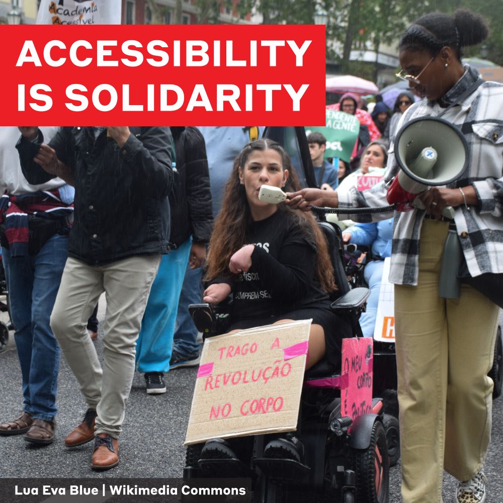 This is a photo of several people at a disability rights march by photographer Lua Eva Blue sourced from Wikimedia Commons. A person using a wheelchair speaks into a megaphone and has a sign that says "Trago a revolução no corpo" which translates to "I bring the revolution on my body." A text overlay says "Accessibility is solidarity"