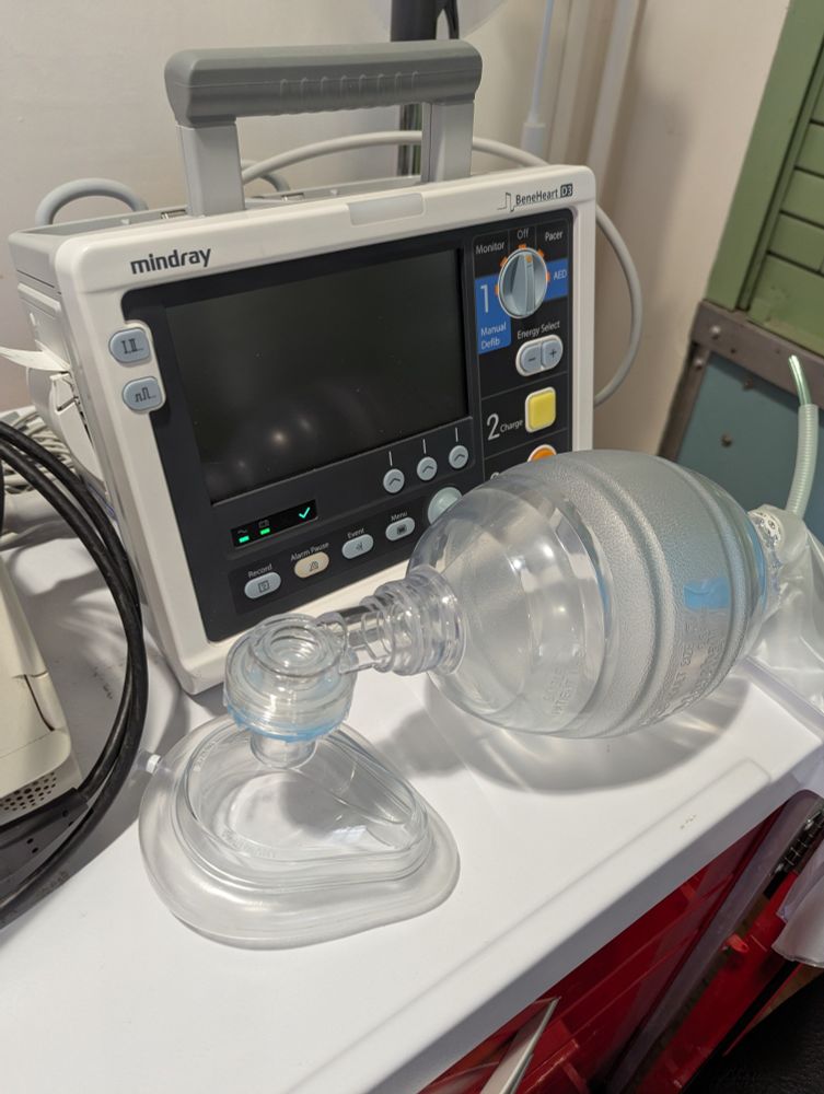 Mindray D3 manual defibrillator/monitor and clear plastic bag-valve-mask atop a red cardiac arrest trolley with a white surface.