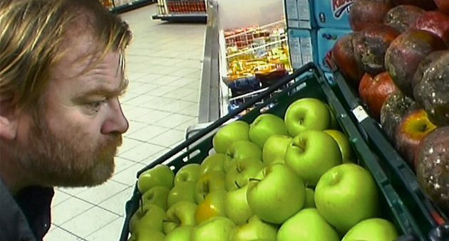 A scruffy man leans close to ripe, green apples one bin below a bin of rotting apples. He’s actor Brendan Gleeson in 28 Days Later