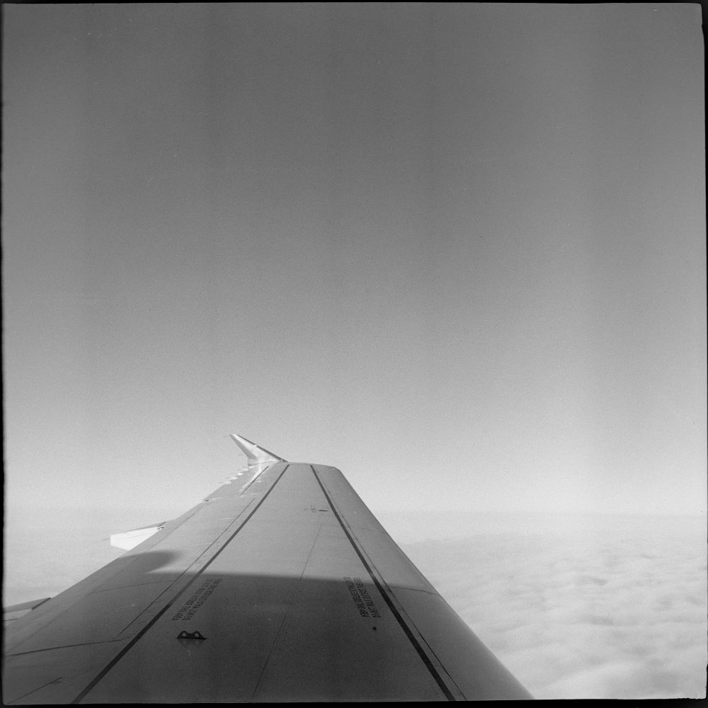Black and white square photo of a passenger aircraft wing, partly in shadow partly in sunlight, clouds below, even sky above. Streaks from too strong agitation in development run vertically down the frame.