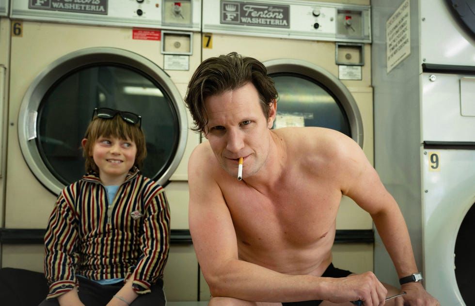 In a laundrymat young boy in a striped shirt (Rafael Mathe) looks admiringly at a shirtless man (Matt Smith) who has a cigarette hanging out of his mount and is looking at the camera in a kind of confrontational or confessional leer.