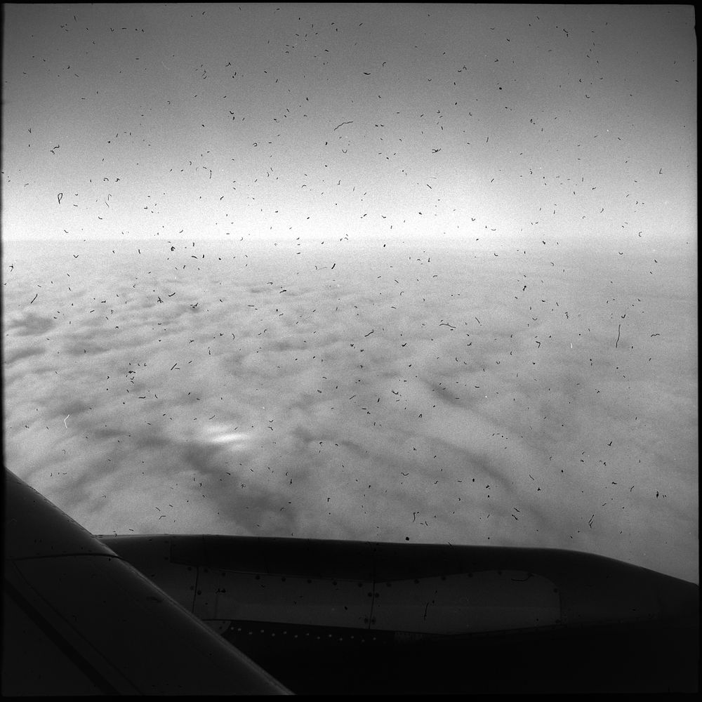 Black and white square photo from inside a passenger aircraft, the engine and wing are in the bottom of the frame, the clouds are visible below, and there are just so many little dusk marks in the emulsion it’s embarrassing. 