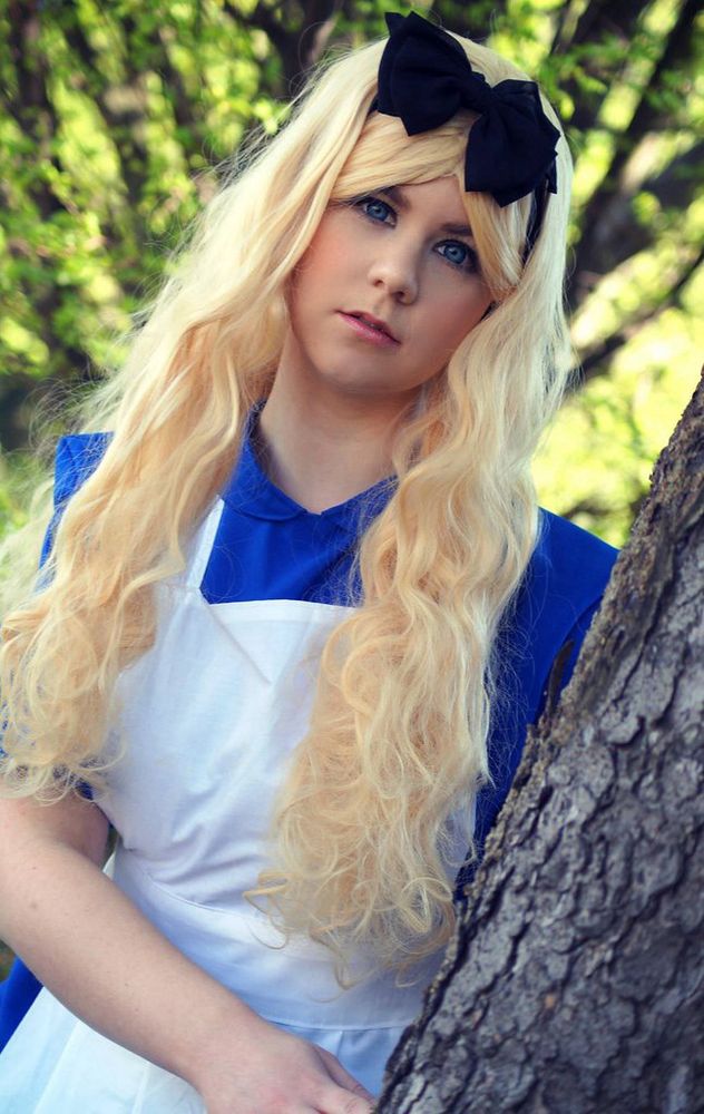 Cosplay Alice in Wonderland - dressed in a blue dress with a white apron, stands behind a tree and peeks out, right hand touches the tree, background is a forest