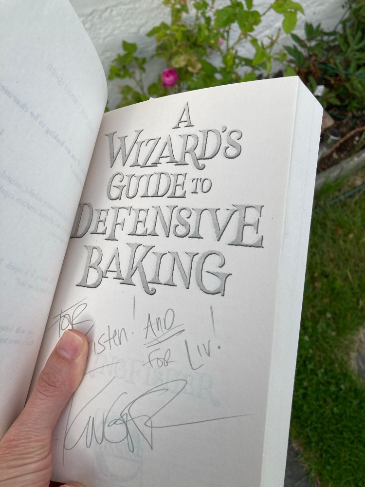 Open book with front page of “a wizard’s guide to defensive baking”, signed by T. Kingfisher, first for the little one and then “_and_ for Liv!”