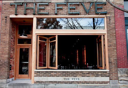 A photograph of a brick storefront with a sign reading THE FEVE printed in tall, angular metal letters atop a new window looking into a dark, bougie restaurant/bar.