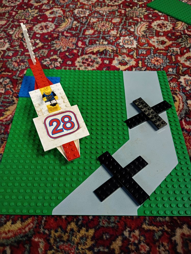 A green Lego baseplate with a gray road in it. There are black Xs of Legos on the road and a very poorly thrown together plane-like contraption with no wings and the number 28 on it.