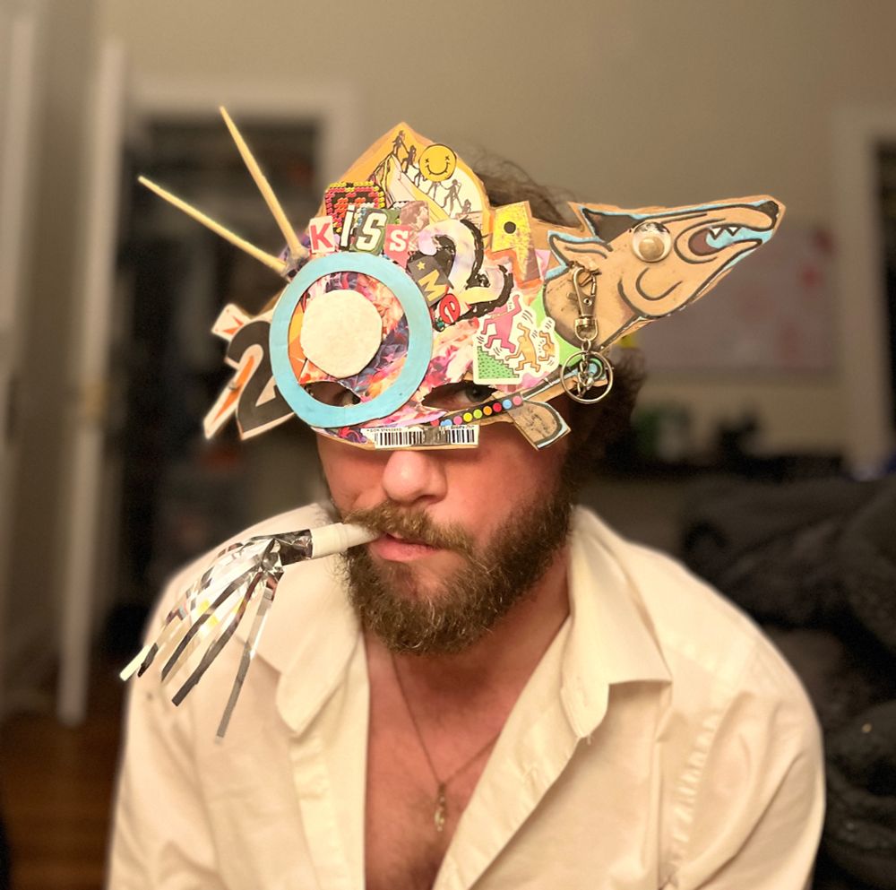white thing with a beard wearing a mask made out of cardboard and collaged elements, reading 2024 and KISS ME. they’re tooting a party horn unenthusiastically.