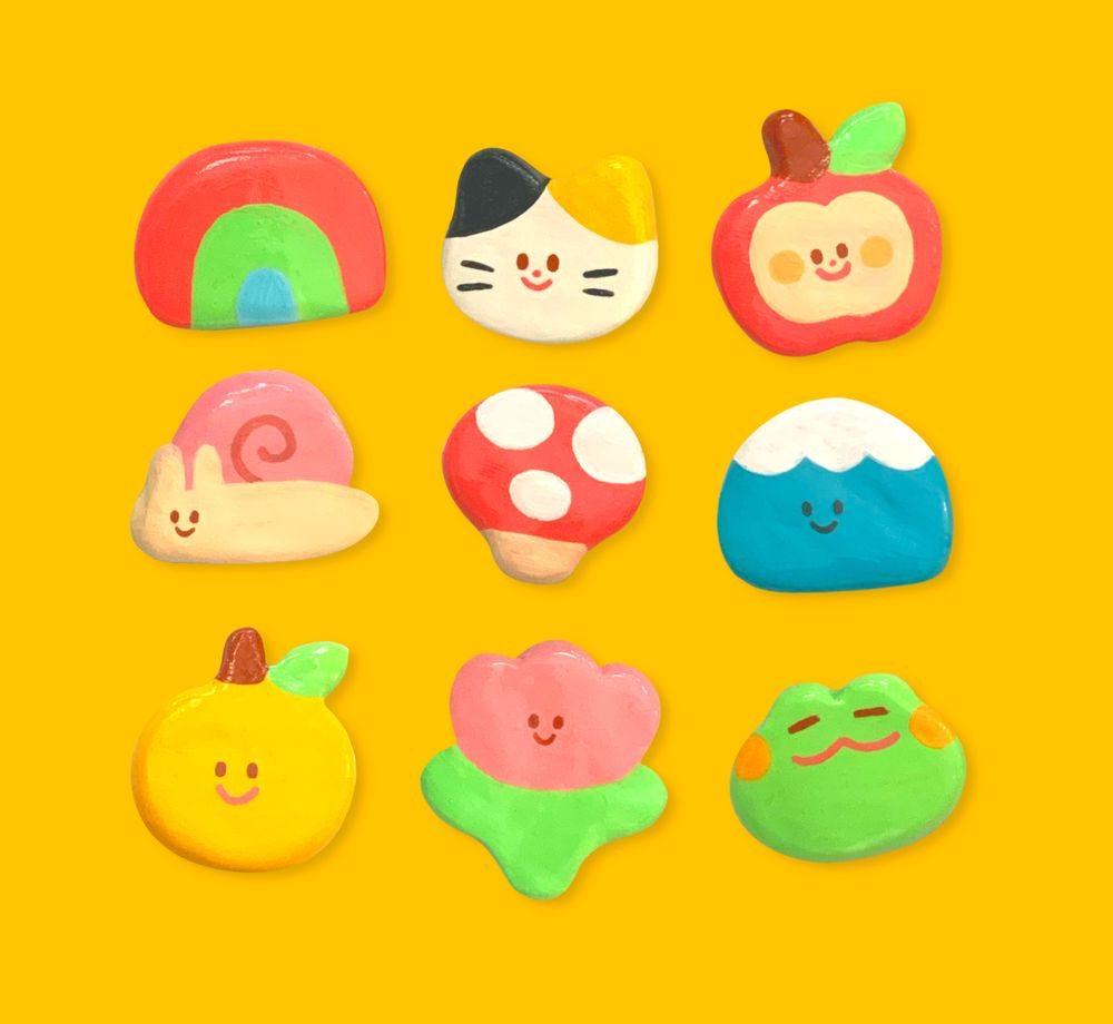 little clay things: a rainbow, a calico cat, an apple, a snail, a mushroom, a mountain, an orange, a tulip, and a frog. the background is a bright yellow.